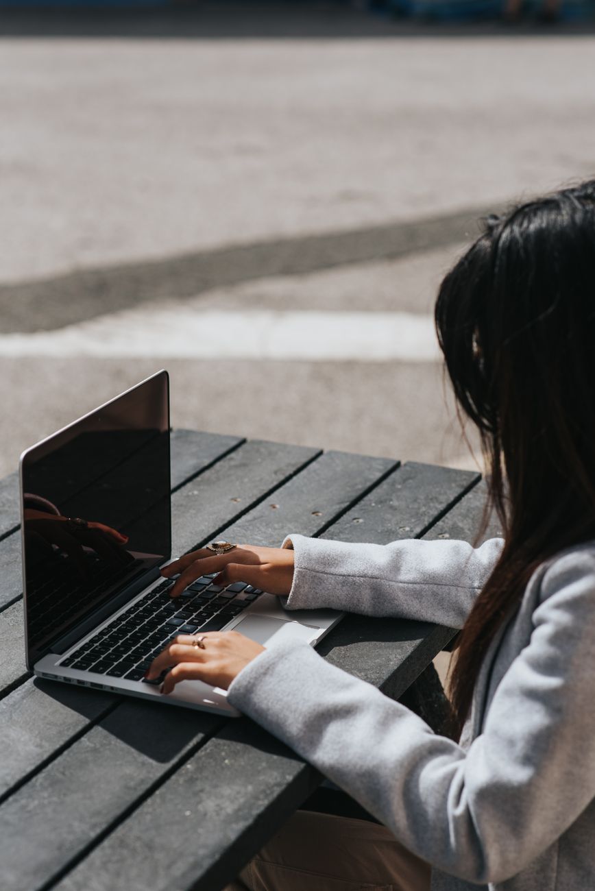 Side view of crop anonymous female distance worker typing on netbook at wooden table on street in sunlight.
