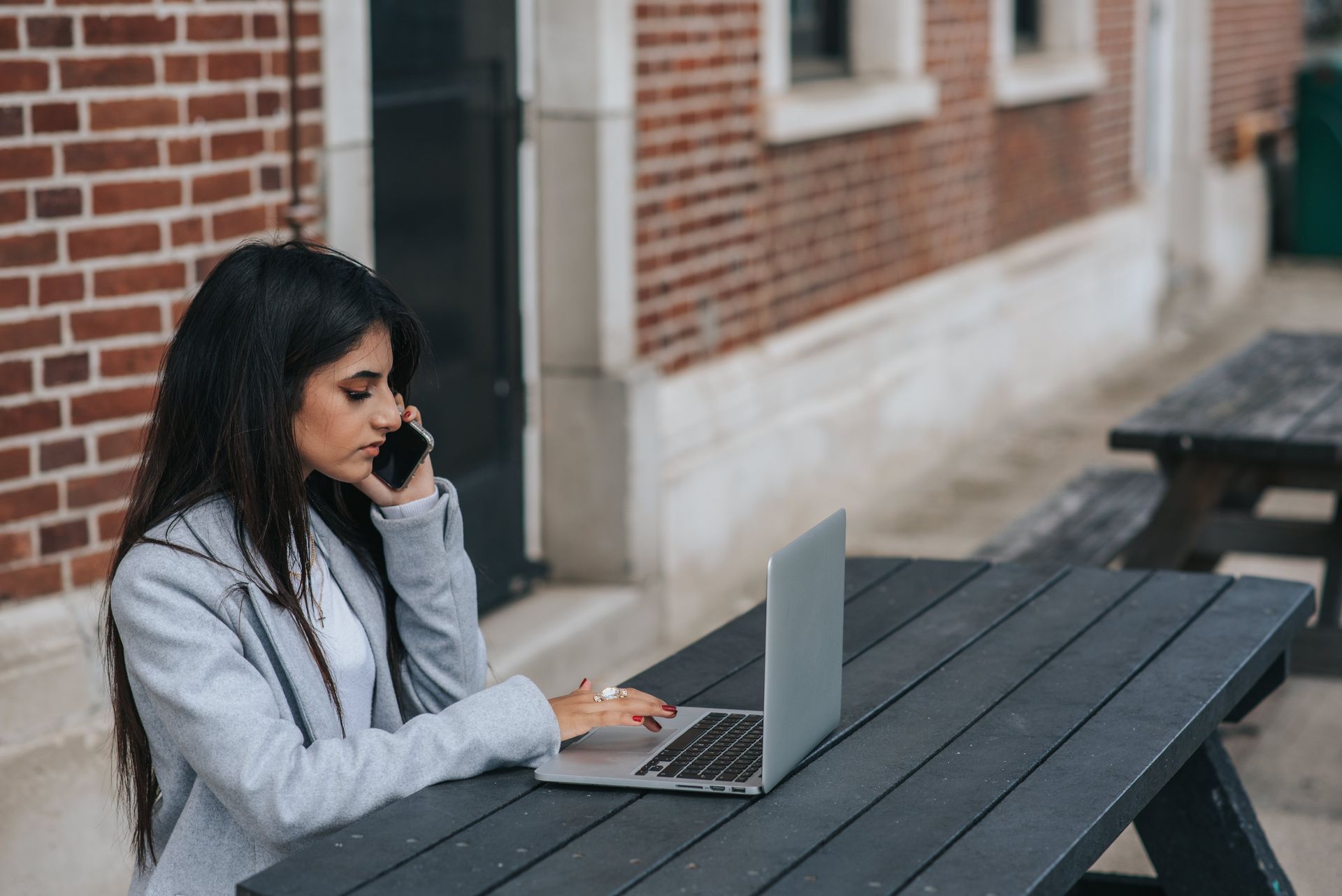 Young woman on a phone call working on her laptop at an outdoor table with a brick wall background.