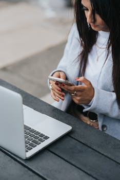 一位职业女性在户外办公桌上查看智能手机，旁边放着笔记本电脑