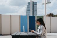 一位年轻女子在户外餐桌上使用笔记本电脑和智能手机，背景是现代城市景观