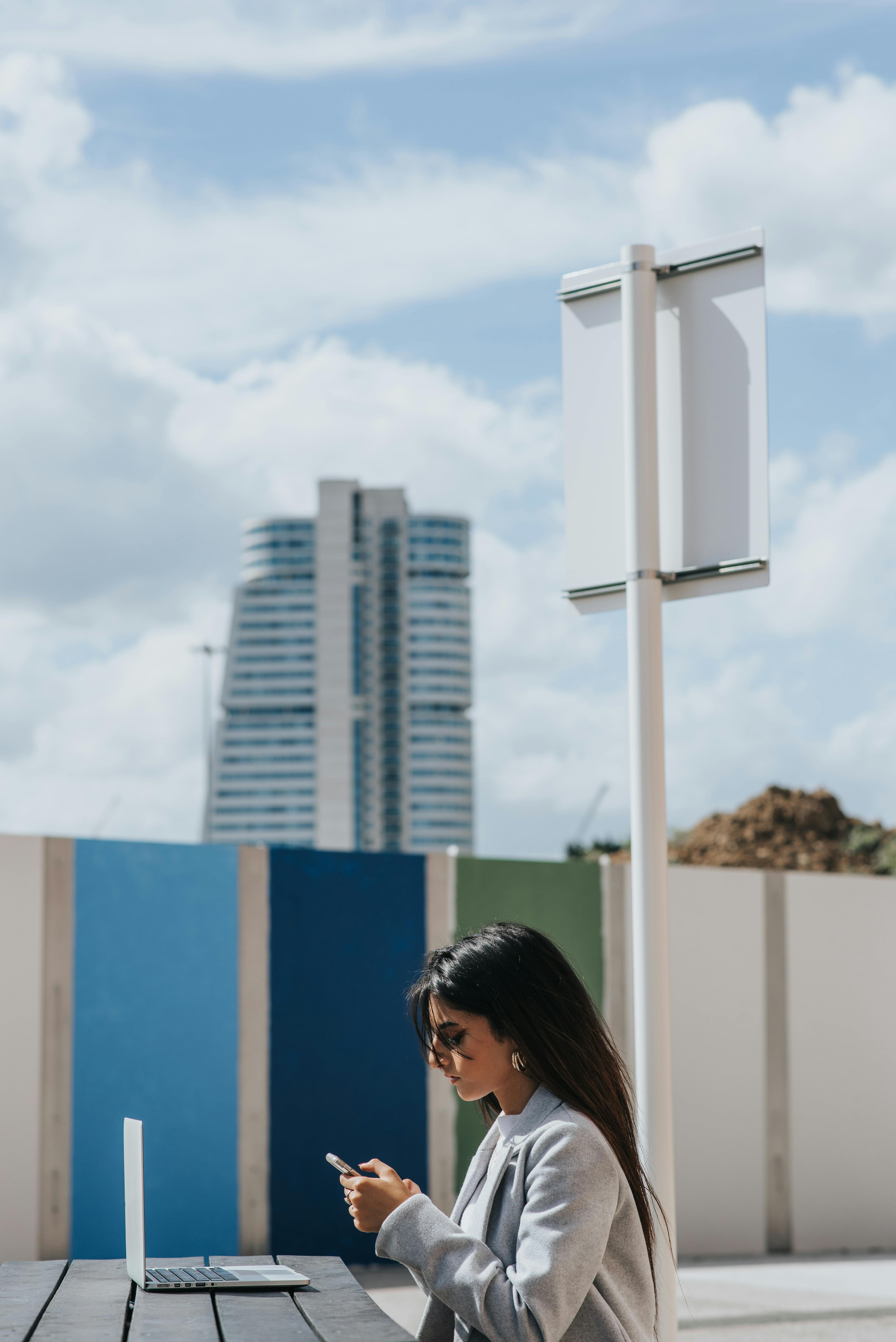 ethnic female entrepreneur surfing internet on smartphone near laptop outdoors