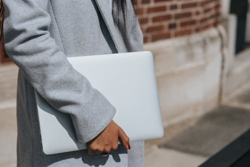 Crop unrecognizable ethnic female entrepreneur in coat standing with netbook in city on sunny day