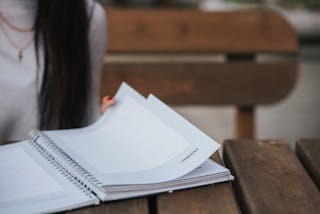 Crop unrecognizable female student with dark hair in white blouse turning pages of notepad at wooden table in park