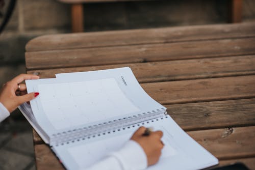 Crop anonymous female student writing in planner while sitting at wooden table in park