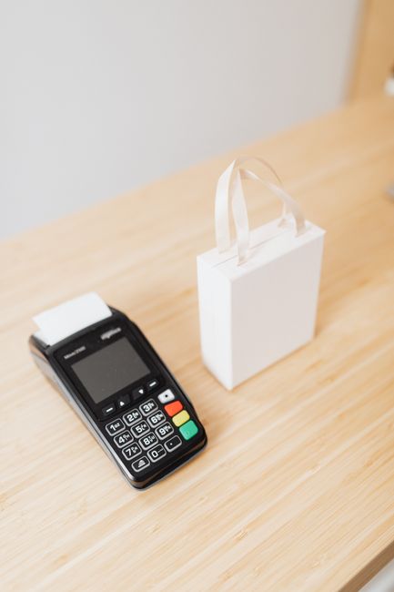 A payment terminal and a small white gift bag on a wooden countertop, ideal for retail or shopping themes. A payment terminal and a small white gift bag on a wooden countertop, ideal for retail or shopping themes.