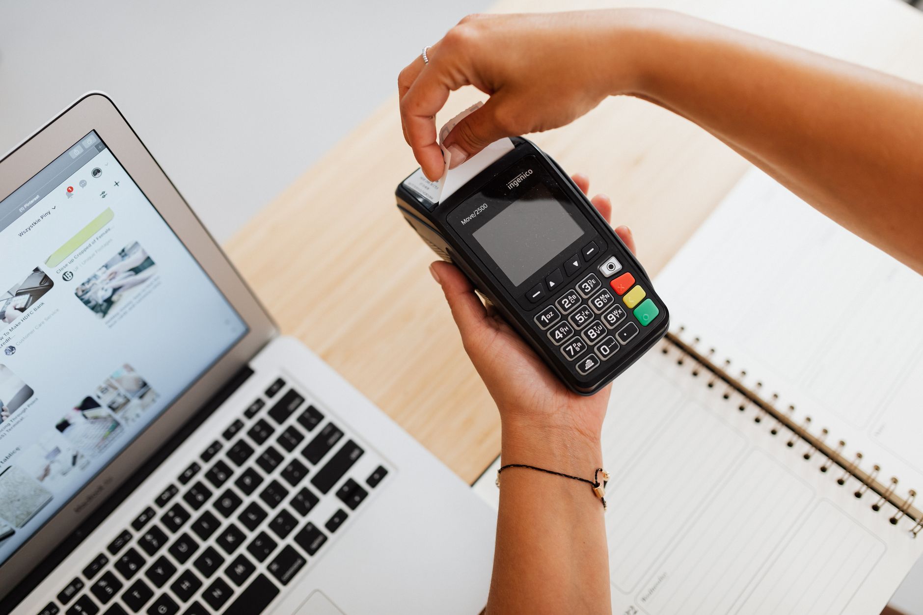Close-up of a person using a card terminal for payment, next to a laptop and notebook.