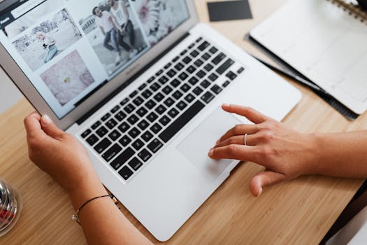 木桌前，一只正在用笔记本电脑浏览照片的手部特写，展现了工作中的互联网使用场景