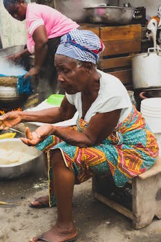 An elderly woman preparing traditional food at an outdoor market. Vibrant cultural scene.