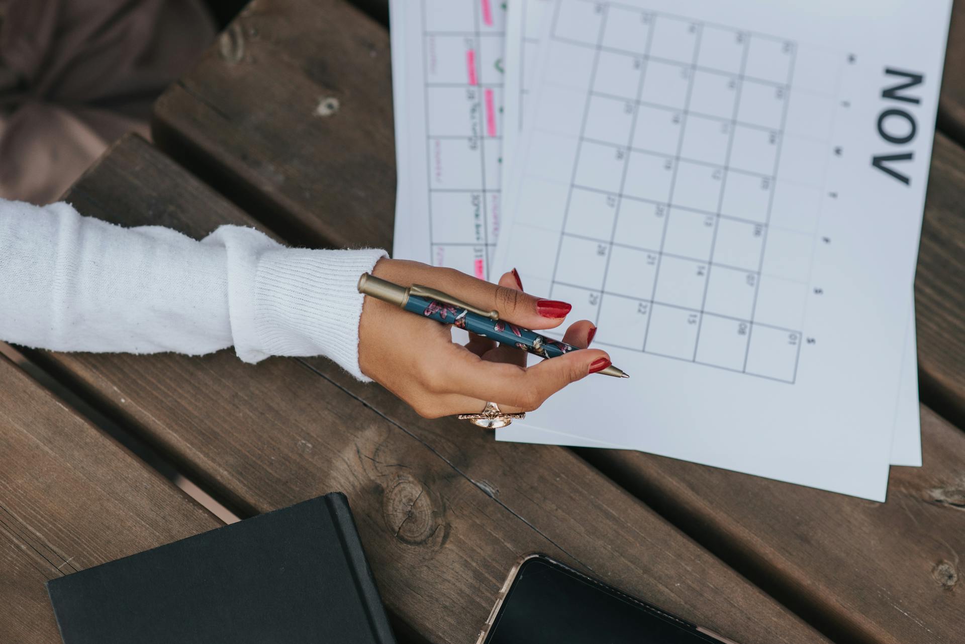 From above of crop anonymous female in white blouse sitting at wooden table with pen and calendar while making schedule