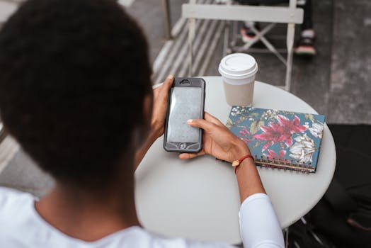 Back view of a woman using a smartphone in a café with a coffee and notebook.