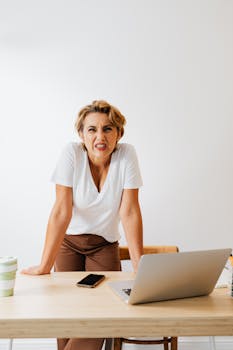 A frustrated woman stands at her office desk with a laptop and phone.