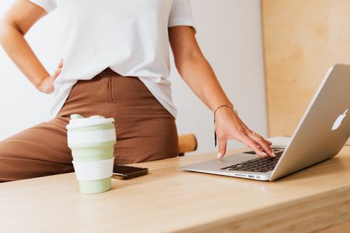 Free A woman stands by a wooden desk using a laptop with a reusable cup nearby. Modern lifestyle concept. Stock Photo