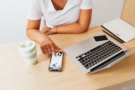 Person working on laptop and smartphone at a minimalist desk with a drink and notebook.