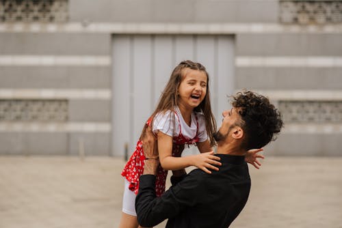 Man carrying smiling little girl in arms and laughing on city street in summer day