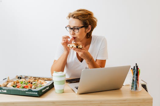 A woman eating pizza at her workspace with a laptop and stationary around.