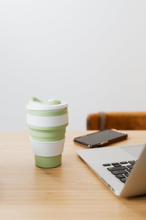Free Reusable Coffee Cup next to Laptop on Desk Stock Photo