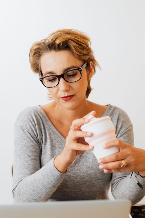 Free Mature woman wearing glasses, concentrating and holding a pink reusable cup while using her laptop indoors. Stock Photo
