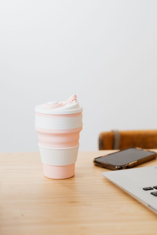 Free A pink reusable coffee cup sits on a wooden desk beside a smartphone and laptop, providing eco-friendly vibes. Stock Photo
