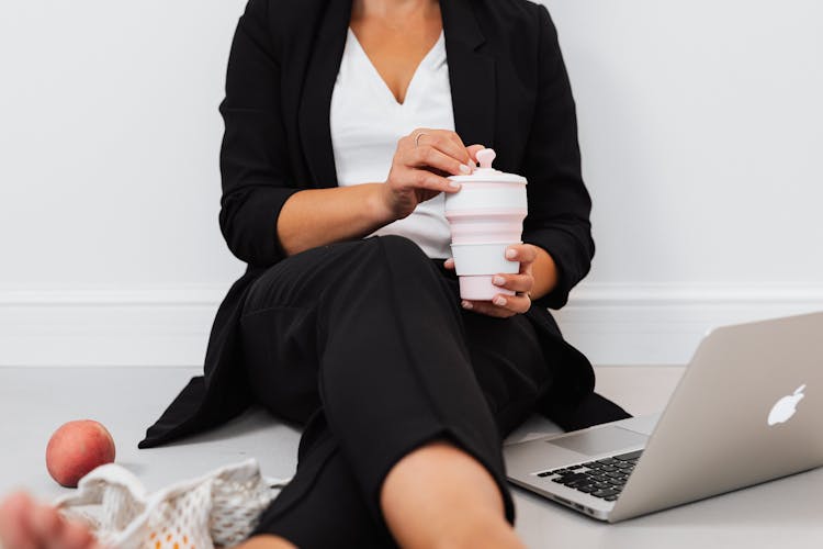 Woman In Black Blazer Holding A Pink Mug While Sitting Beside Her Laptop