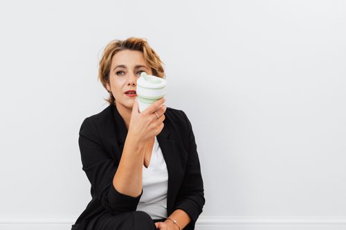 Free Stylish woman in black blazer holding a collapsible cup, looking thoughtful indoors. Stock Photo