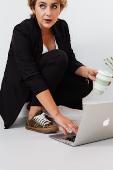 Professional woman in modern office setting using laptop while holding reusable coffee cup.