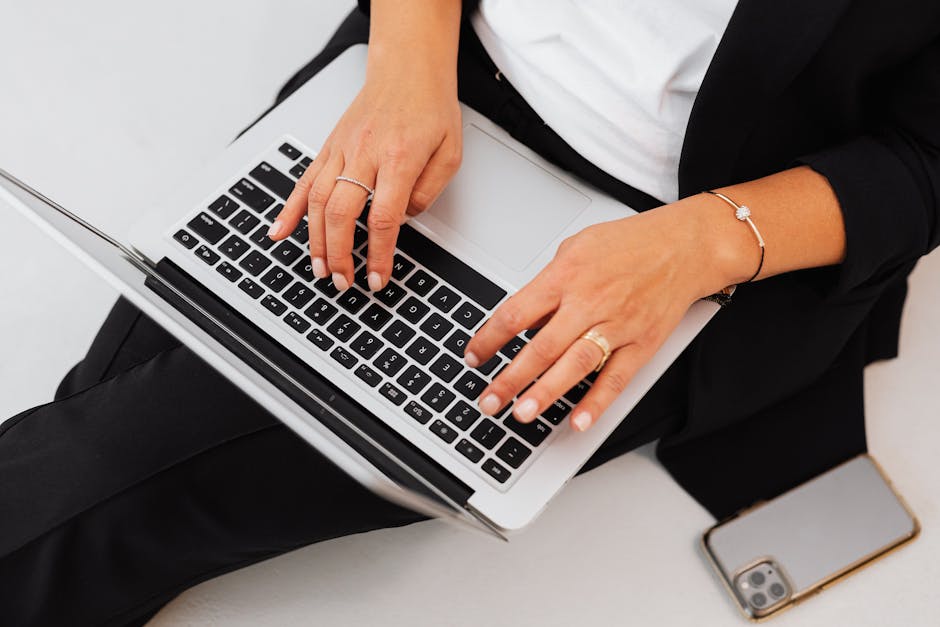 A woman in business attire types on a laptop with a smartphone beside her, emphasizing modern technology and work.