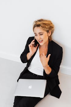 Confident businesswoman in black blazer talking on phone while sitting with laptop indoors.