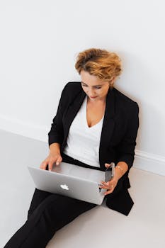 Businesswoman sitting on floor using laptop and smartphone, multitasking.