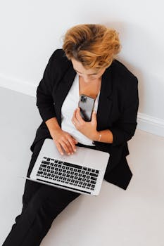 High angle view of a businesswoman sitting on the floor using a laptop and smartphone indoors.