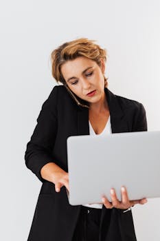Businesswoman in a black blazer multitasking with a laptop and phone, showcasing modern corporate lifestyle.