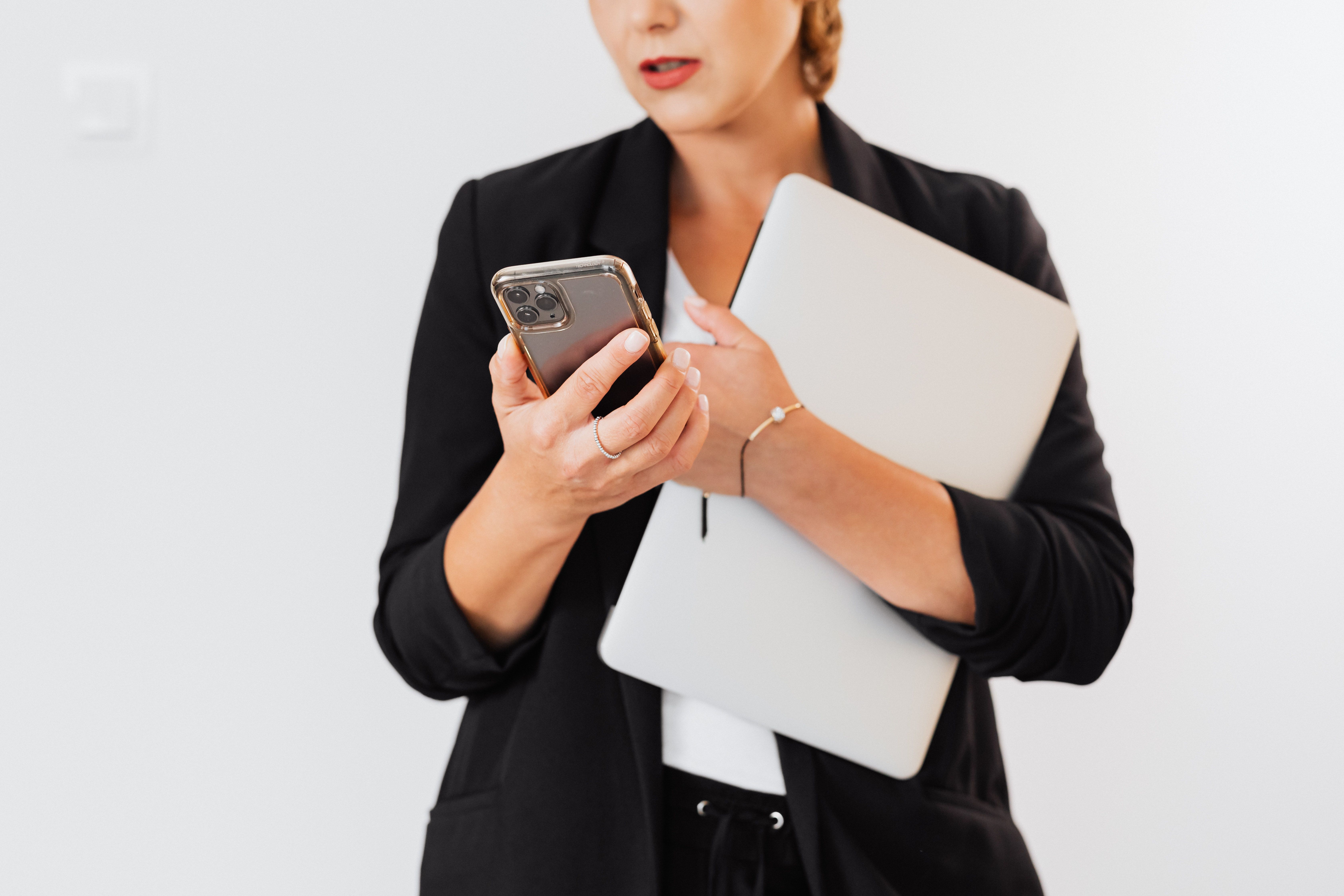 A businesswoman holds a laptop and checks her smartphone while standing indoors