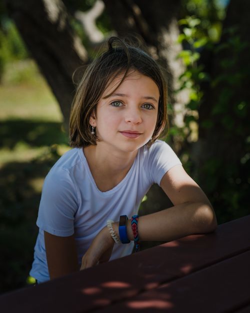 Free Serious girl in casual t shirt sitting at table near green tree and grass in summer sunny day in backyard and looking at camera Stock Photo