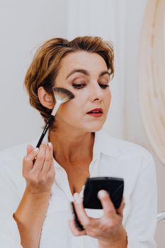 Woman using makeup brush in a well-lit indoor setting, enhancing her facial features.