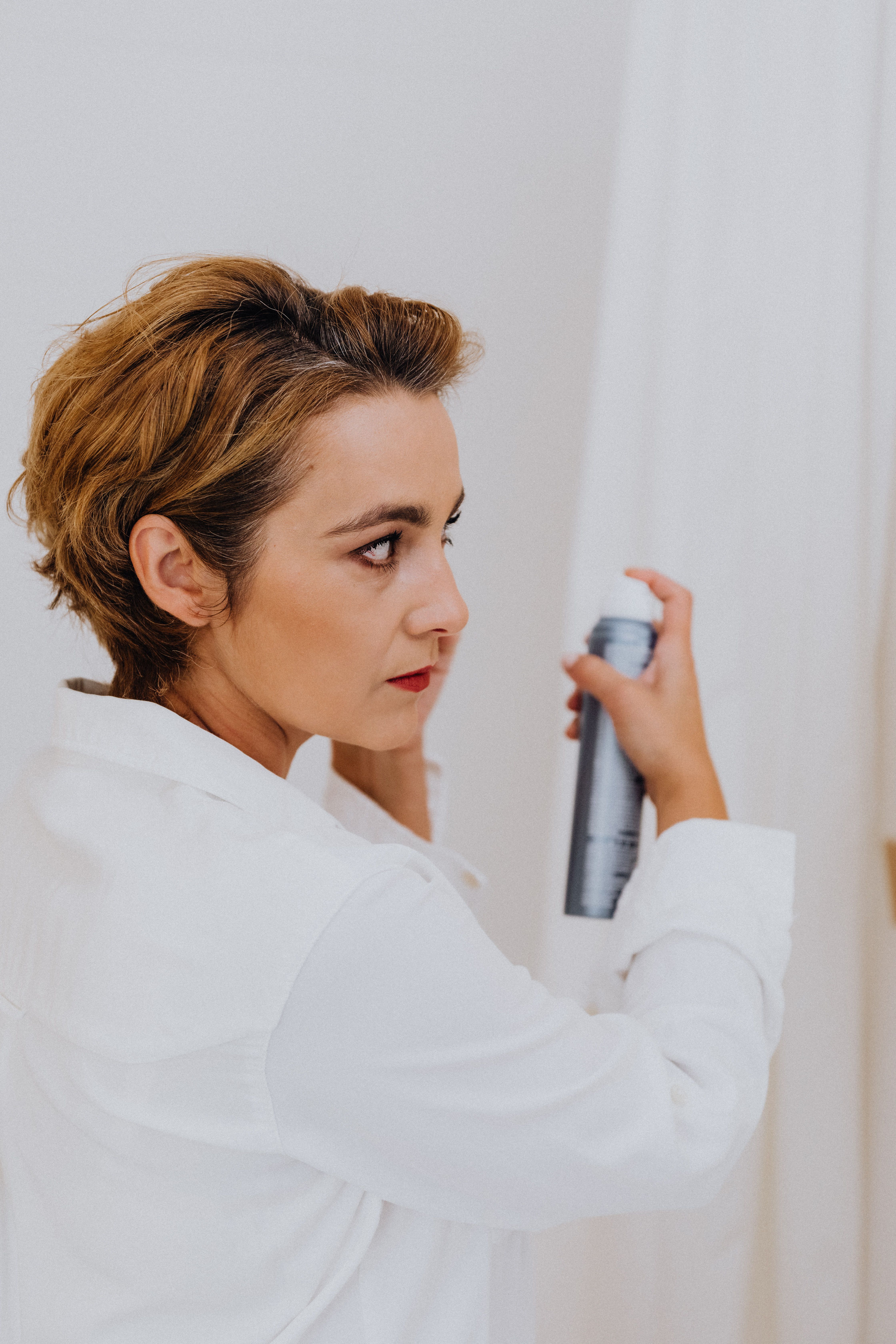 Woman in white shirt styling hair with spray. Indoor setting, modern elegance.