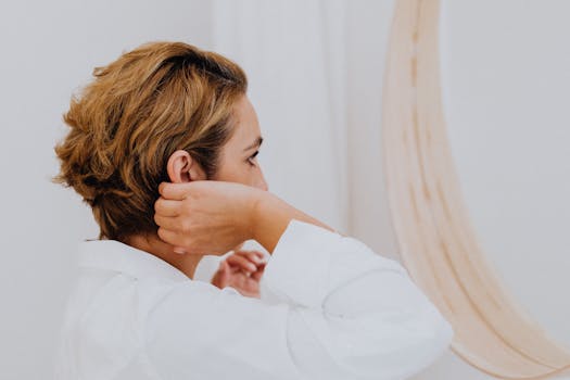 Woman adjusts hair in front of a mirror wearing a white shirt, displaying short hairstyle.