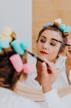A woman with curlers applies eye makeup in front of a mirror, showcasing beauty routine.