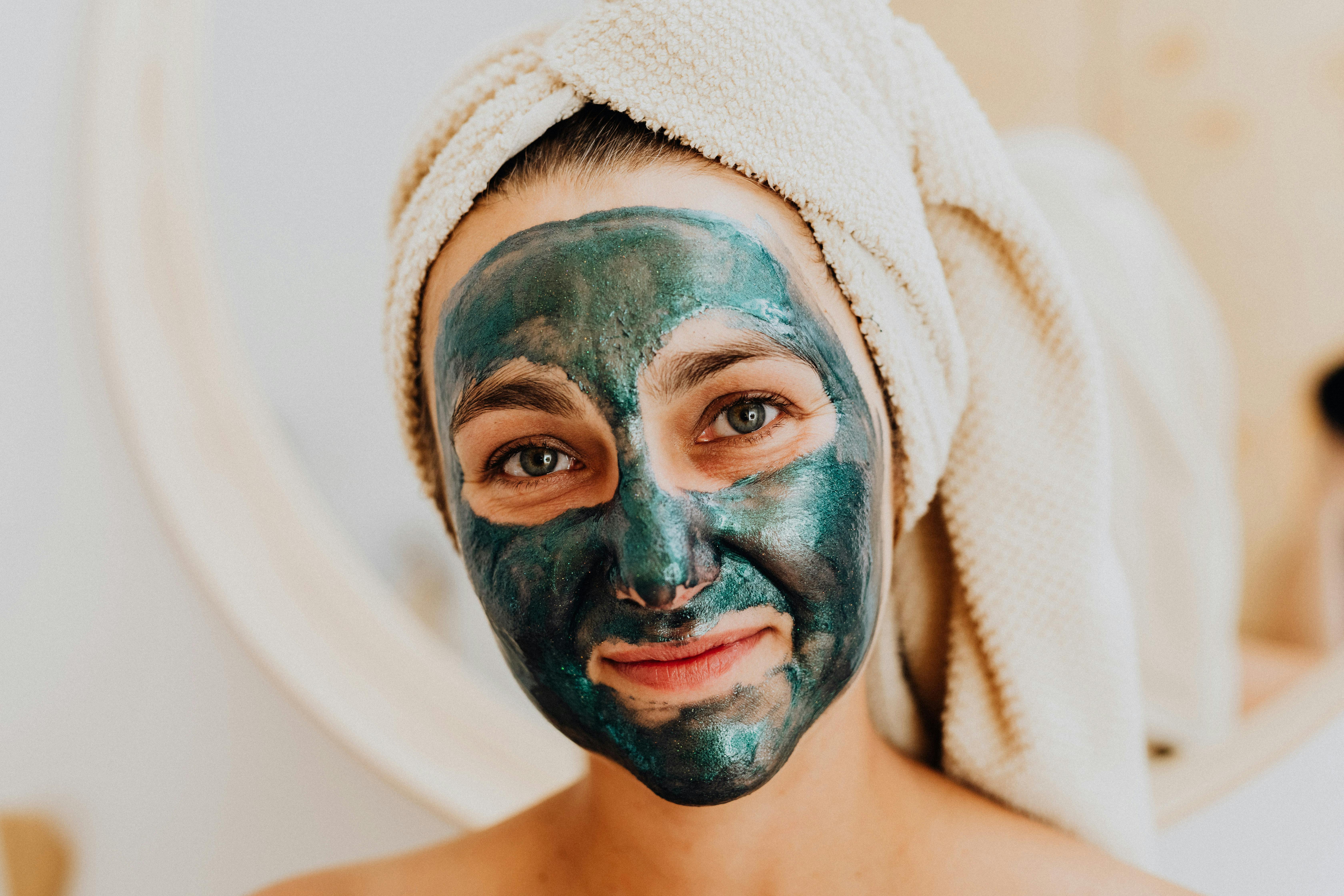 Happy woman wearing a cosmetic face mask and a towel on her head, enjoying self-care indoors.