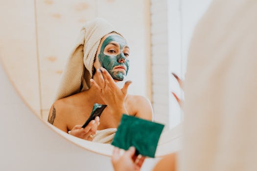 A woman applies a green clay face mask in a bathroom mirror, promoting self-care and skincare routines.