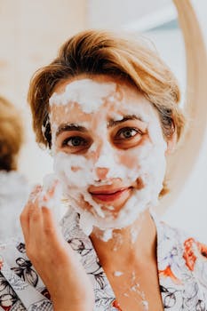 Woman enjoying a foamy face mask for skincare routine, smiling in a bathroom setting.