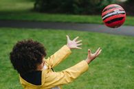 Side view of playful African American kid playing with red ball while standing on green lawn in park on blurred background