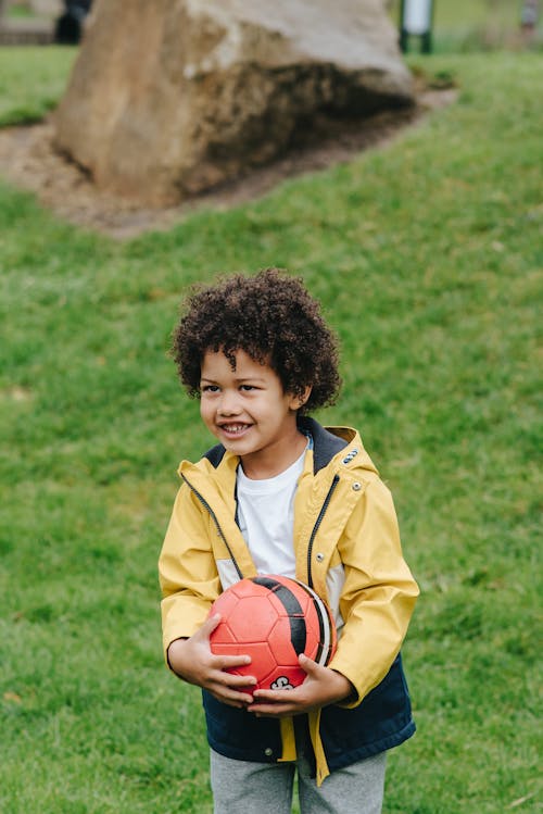 Delighted African America kid with Afro hair looking away while standing on green meadow with football in hands