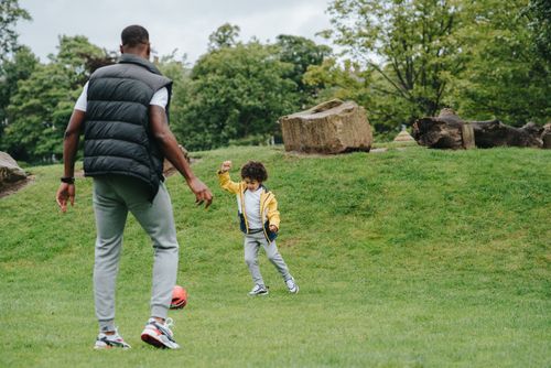Free Full body of cheerful African American kid and unrecognizable black male parent playing with ball on grassy ground in park Stock Photo