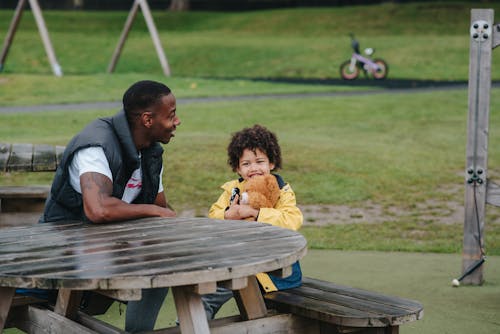 Positive African American man and little kid with plush bear sitting at wooden table on grassy playground on blurred background