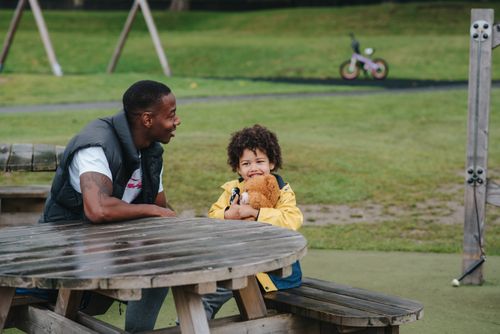 Free Positive African American man and little kid with plush bear sitting at wooden table on grassy playground on blurred background Stock Photo