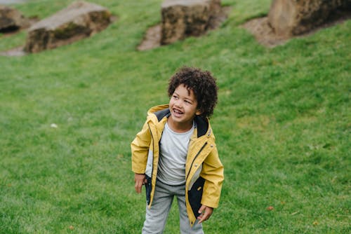 Positive African America little kid smiling and leaning forward while standing on grassy ground with stones on background while