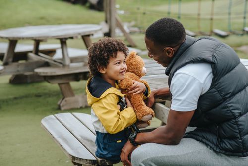 Free Side view of African American son with plush bear and father looking at each other while sitting at wooden table in park on blurred background Stock Photo