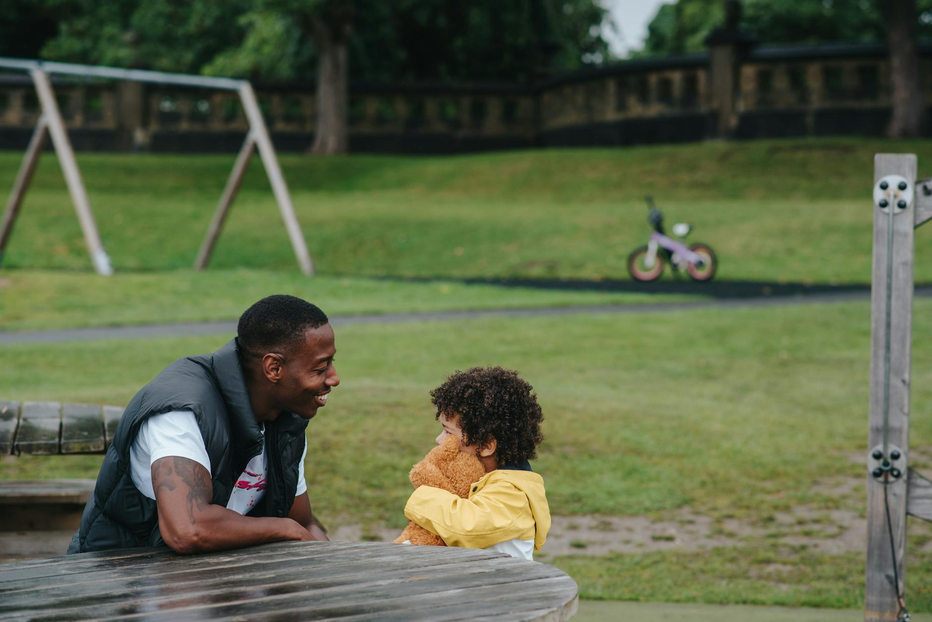 Side view of African American little boy with toy and male parent looking at each other while sitting at wooden table on playground on blurred background