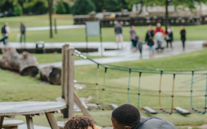 Side view of little black kid with toy and African American male sitting on wooden bench on playground on blurred background