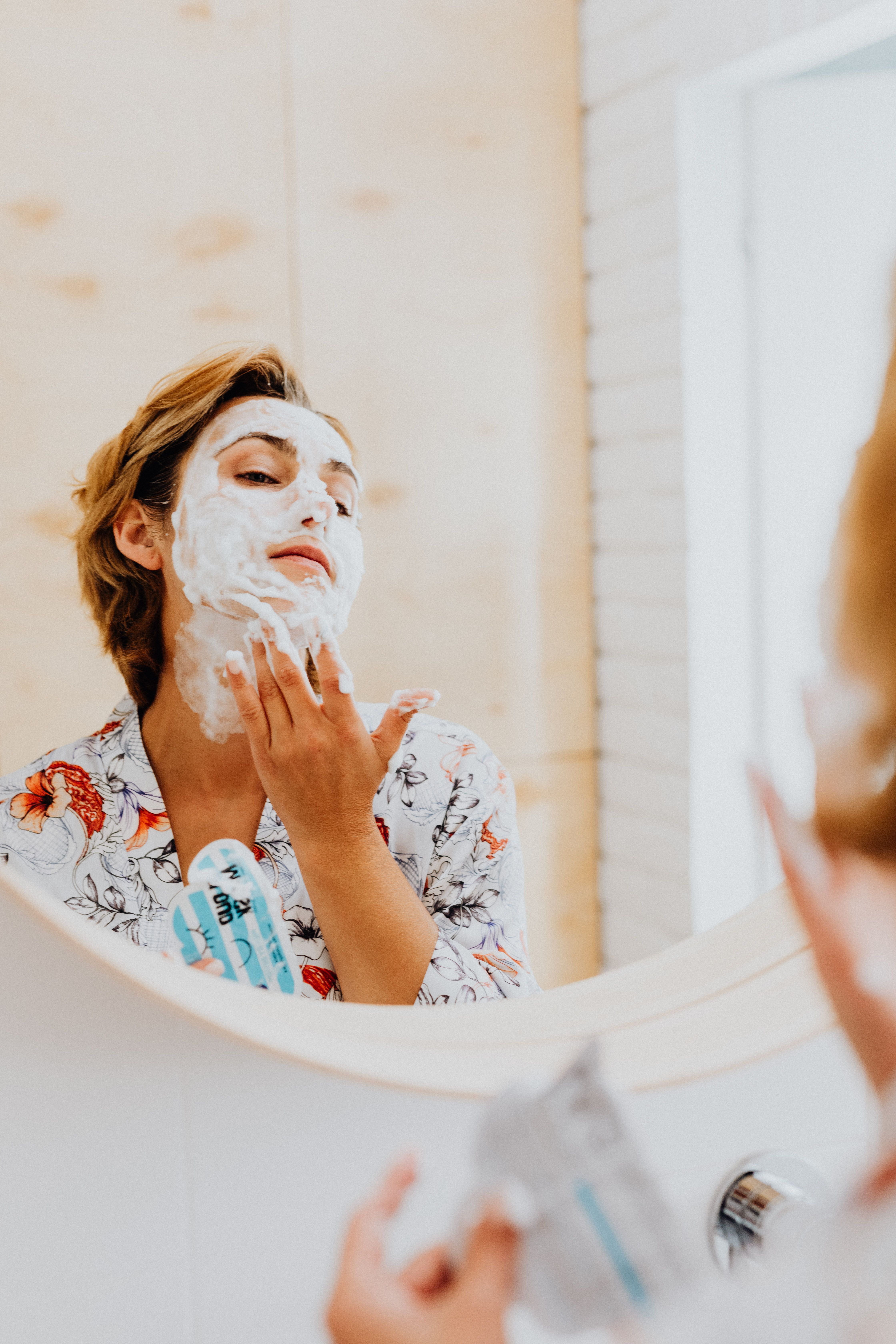 Woman applying face mask with foam in front of the bathroom mirror for a skincare routine.