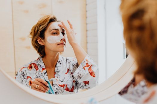 Adult woman applies facial mask while looking in bathroom mirror, promoting skincare routine.
